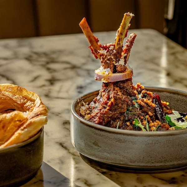 A bowl of lamb shank curry with bone-in lamb, onion, and a side of flaky paratha.